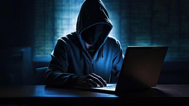 Hacker in hoodie using laptop in dark room, symbolizing cyber threats.